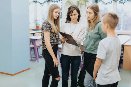Elegant teacher stands with a black pad and explains something to a group of teenagers during...