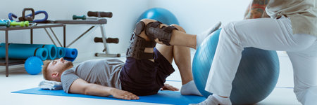 Teenage girl after car accident with orthosis on the leg exercises on blue mat in the company of physiotherapistの写真素材