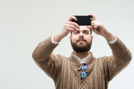 Isolated studio portrait of handsome young adult with beard taking photo with his smartphoneの写真素材