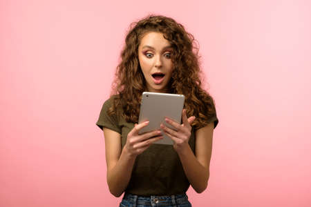 Closeup shot of pretty woman with long curly hair holding tablet and looking on its screen with...