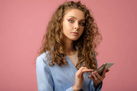 Closeup studio portrait of pretty businesswoman with long curly hair holding smartphone with finger on screen isolated on pink background in studio.の写真素材