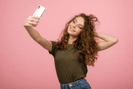 Closeup shot in studio of pretty student with long curly hair posing with her smartphone and making...