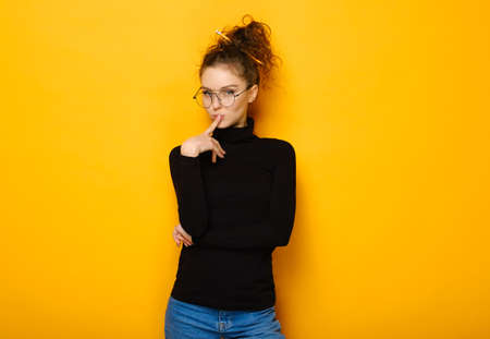 Portrait of pretty teenage girl with curly hair wearing glasses and casual black turtleneck sweater isolated on yellow background in studioの写真素材