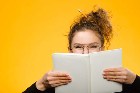 Closeup shot of attractive and charismatic teenage girl with curly hair wearing glasses. Model hiding behind white notebook with positive emotionの写真素材