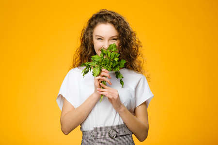 Closeup portrait of pretty caucasian girl with curly hair holding green fresh parsley isolated on colorful yellow backgroundの写真素材
