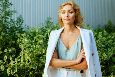 Closeup portrait of happy businesswoman with pretty smile. Model wearing beautiful white suit and posing with hands crossed in a confident pose outdoors on a sunny dayの写真素材