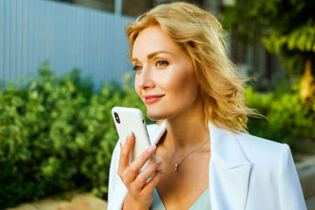 Closeup outdoor portrait of pretty businesswoman holding smartphone and having conversation over speakerphone. Model has blond hair and wearing beautiful white suit.の写真素材