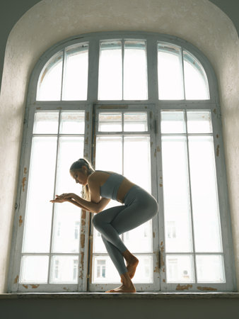 Fitness, yoga, stretching and healthy lifestyle concept - young woman doing exercises near window at...