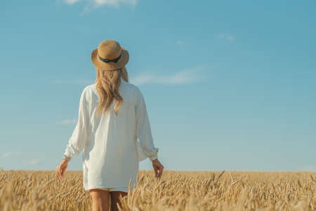 Back view of young woman in a white dress and a straw hat on a wheat field in the summer. The...