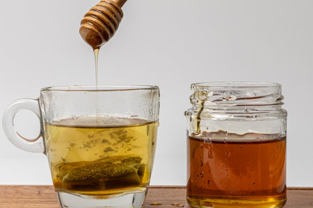 Honey dripping from a dipper in a cup of green tea kept beside a jar of honeyの写真素材
