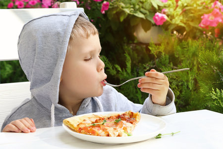 Pretty toddler is holding fork with a slice of pizza The look of pleasure on his face.Beautiful floowers and summersunshine on the background.Open cafe interior.の写真素材