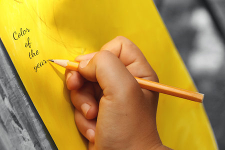 Child holds pencil and writing on the yellow paper with grey background.Trendy colors of the 2021.