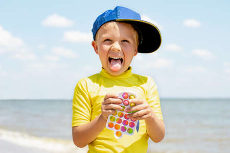 Funny child showing his tongue and playing with new sensory toy pop it on the beach. Silicone simple dimple in the plastic frame. Antistress toy for children and adults.の写真素材