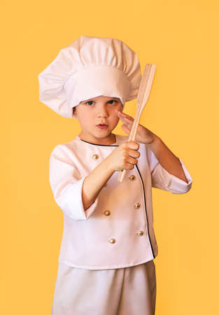 Smiling child in white chef uniform,holding wooden spatula in hands. Cheerful yellow background.の写真素材
