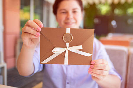 Closeup photography of woman,holding a bronze invitation envelope with wax seal and satin ribbon.Sunlight on backgroundの写真素材