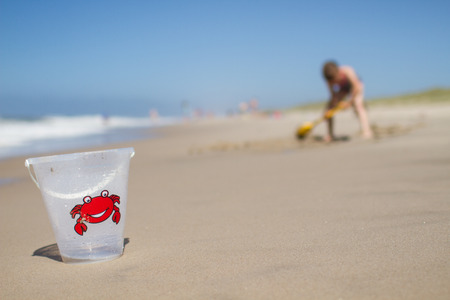 Focus is on plastic bucket with red crab illustration. Blurred background with young girl digging in the sand. Girl is wearing multi-colored swim suit.の写真素材