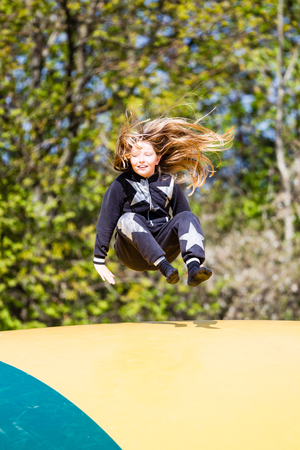 Smiling young girl jumping up and down on bouncing trampoline pad on sunny day at playground.の写真素材