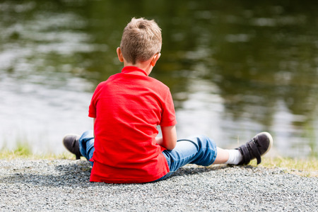 Lonely boy sitting by himself and thinking about life near a small lake on a summer day.の写真素材