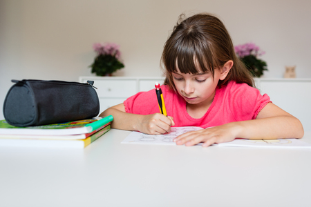 Young white girl writing with her pencil and doing her elementary school homework while sitting at white table.の写真素材