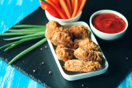 Chicken wings in breading with sliced vegetables and tomato sauce on black plate and blue table