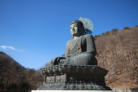 A ancient metal carving of sitting peace buddha in front of tree mountain and painted with green color at large historical temple at south koreaの写真素材