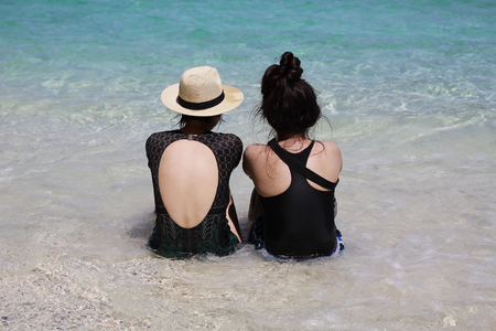 Portrait of two young asian female friends sitting on the beach shore turn back at camera laughing. Multiracial young women strolling along a sea, friendship backgroundsの写真素材
