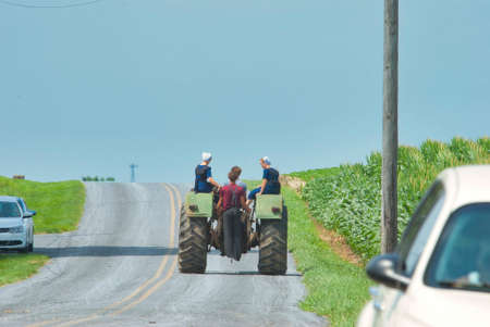 Ronks, Pennsylvania, July 2021 - Amish Boy and Girl Teenagers Riding on a Old Tractor Thru Farmlands...