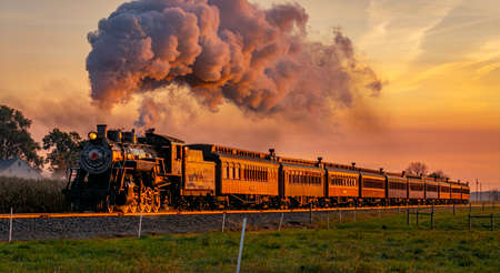 A View of an Antique Steam Passenger Train Approaching at Sunrise With a Full Head of Steam and Smoke Traveling Thru Farmlandsの写真素材