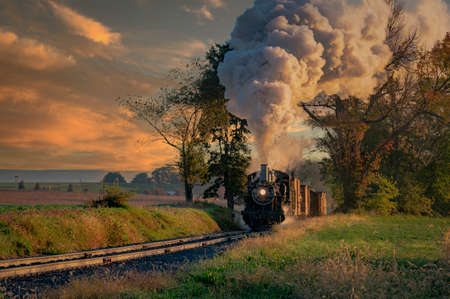 A View of an Antique Freight Steam Train Blowing Smoke Approaching Thru Trees in Late Afternoonの写真素材