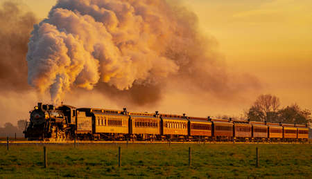 A View of an Antique Steam Passenger Train Approaching at Sunrise With a Full Head of Steam and Smoke Traveling Thru Farmlandsの写真素材