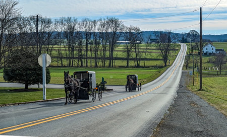 A View of Two Amish Horse and Buggies Traveling Down a Countryside Road Thru Farmlands and a One Room School House on a Sunny December Dayの写真素材