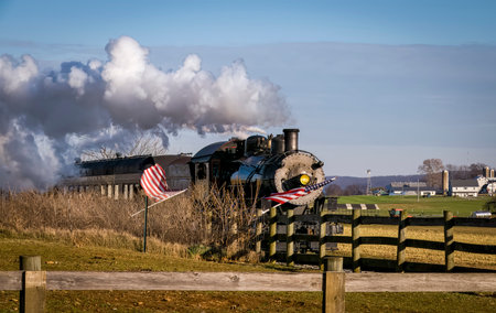 A View of a Classic Steam Passenger Train Approaching, With American Flags Attached to a Fence on a Sunny Summer Dayの写真素材