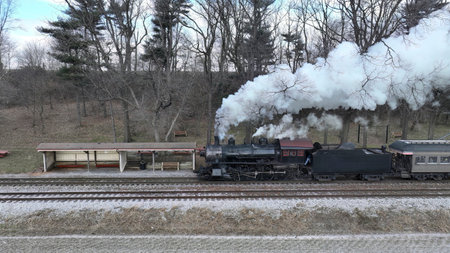 A Side Drone View of a Steam Locomotive Approaching Traveling Thru Fields and Meadows, Blowing White Smoke on a Winter Dayの写真素材
