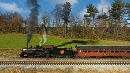 Ronks, Pennsylvania, November 26, 2022 - A Drone View of a Restored Steam Passenger Train Traveling Thru Farmlands Pulling Up to a Small Station on an Autumn Dayのeditorial素材