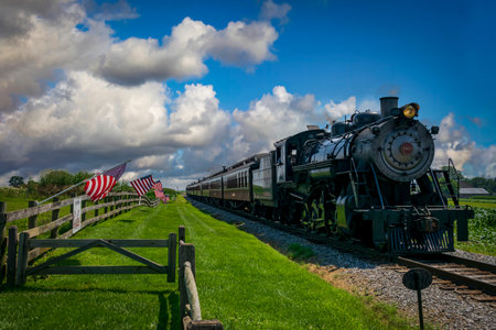 Ronks, Pennsylvania, July 19, 2022 - A Restored Antique Steam Passenger Train Approaches a Fence With Gently Waving American Flags on It On a Sunny Summer Dayのeditorial素材