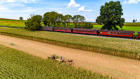 Ronks, Pennsylvania, September 11, 2021 - An Aerial View of Six Horses Pulling an Amish Harvesting Corn Stalk Machine, as Steam Passenger Train Travelsのeditorial素材