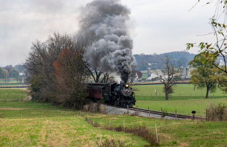 Ronks, Pennsylvania, November 4, 2023 - View of an Antique Steam Passenger Train Approaching Coming Out of a Group of Trees, Blowing Smoke on a Fall Dayのeditorial素材
