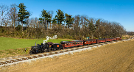 Ronks, Pennsylvania, November 26, 2022 - A Drone View of a Restored Steam Passenger Train Traveling Thru Farmlands Pulling Up to a Small Station on an Autumn Dayのeditorial素材