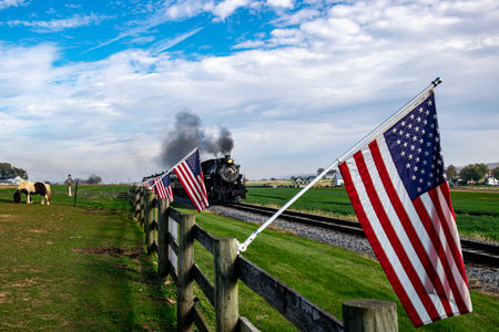 A vintage steam train chugs along the countryside, flanked by American flags on a wooden fence,...