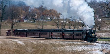A steam train is traveling down a snowy track. The train is black and is surrounded by trees. The...