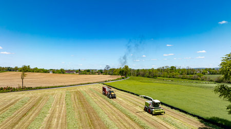 An aerial shot displaying intense harvesting activity with machinery in action across a multi-textured landscape of cut and uncut fields, under a bright blue sky with distant smoke visible.の写真素材