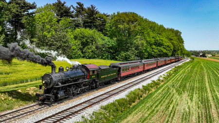 A classic Norfolk and Western steam locomotive hauls vintage passenger cars through a picturesque rural setting. The train, belching thick black smoke, travels alongside vibrant green fieldsのeditorial素材