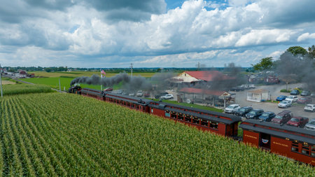 Ronks, Pennsylvania, August 15, 2023 - Clouds gather above as a historic steam train runs parallel to lush cornfields, offering a journey through time by the rural landscape.のeditorial素材