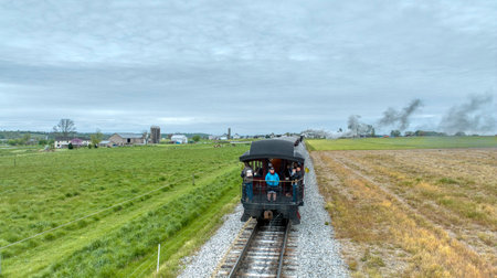 Ronks, Pennsylvania, USA, April 27, 2024 - A serene journey captured from the rear of a train, showing passengers enjoying the ride through a picturesque rural landscape with farmhouses and lush fields under a cloudy sky.のeditorial素材