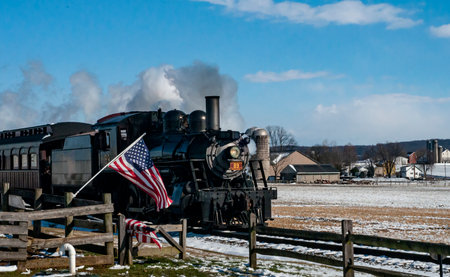 Ronks, Pennsylvania, USA, February 17, 2024 - A steam train is pulling a train car with an American flag on it. The train is traveling through a snowy fieldのeditorial素材