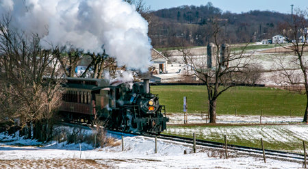 Ronks, Pennsylvania, USA, February 17, 2024 - A steam train is traveling through a snowy field. The steam train is black and white and is surrounded by trees. The train is moving through the snow, leaving a trail of smoke behind itのeditorial素材