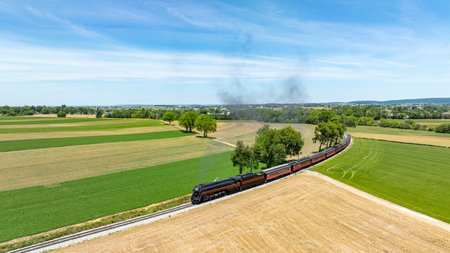 A captivating aerial shot of a steam train gliding through a green rural panorama, weaving between fields and emitting a trail of smoke against a clear skyの写真素材