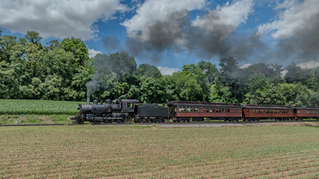 A Black Steam Locomotive Pulling Red Passenger Cars Passes Through A Verdant Countryside With Lush Green Trees In The Background, Billowing Smoke Against A Partly Cloudy Sky.の写真素材