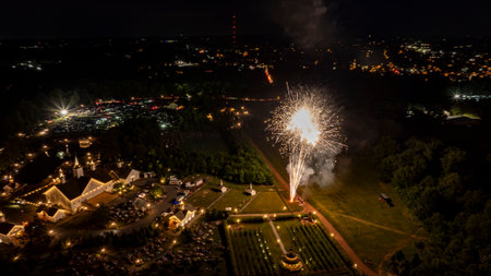 Bright White Fireworks Burst Over A Well-Lit Estate, Surrounded By Trees And Fields, With Distant City Lights Illuminating The Horizon, Creating A Festive Nighttime Celebration Scene.の写真素材