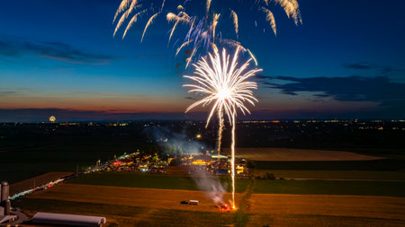 White Firework Burst Illuminating The Night Sky Over A Farm, With A Distant Crowd And City Lights At...
